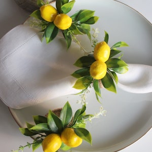 Pu&ograve; includere: Tovaglioli di lino bianco disposti su un piatto, ognuno adornato con un anello portatovagliolo a tema limone. Gli anelli presentano limoni gialli, foglie verdi e piccoli fiori bianchi, creando un'estetica fresca ed estiva. Il piatto ha un bordo dorato.