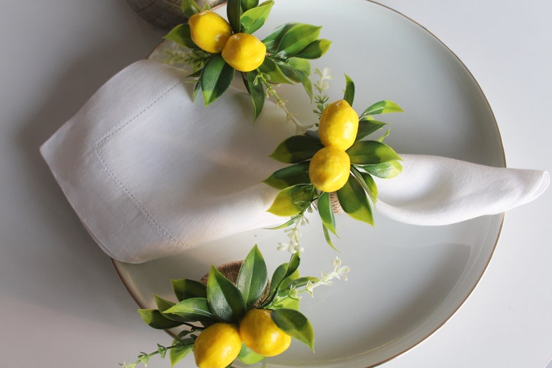 Pu&ograve; includere: Tovaglioli di lino bianco disposti su un piatto, ognuno adornato con un anello portatovagliolo a tema limone. Gli anelli presentano limoni gialli, foglie verdi e piccoli fiori bianchi, creando un'estetica fresca ed estiva. Il piatto ha un bordo dorato.