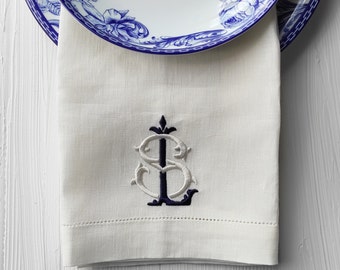 May include: A white linen napkin with a navy blue and white embroidered monogram. The monogram is a stylized "S" with a decorative flourish.