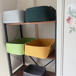 May include: Five rectangular storage baskets made of coiled rope, each featuring a small leather-like side tag. The baskets are colored light gray, dark green, lime green, mustard yellow, and charcoal gray, arranged on a dark wood shelving unit.