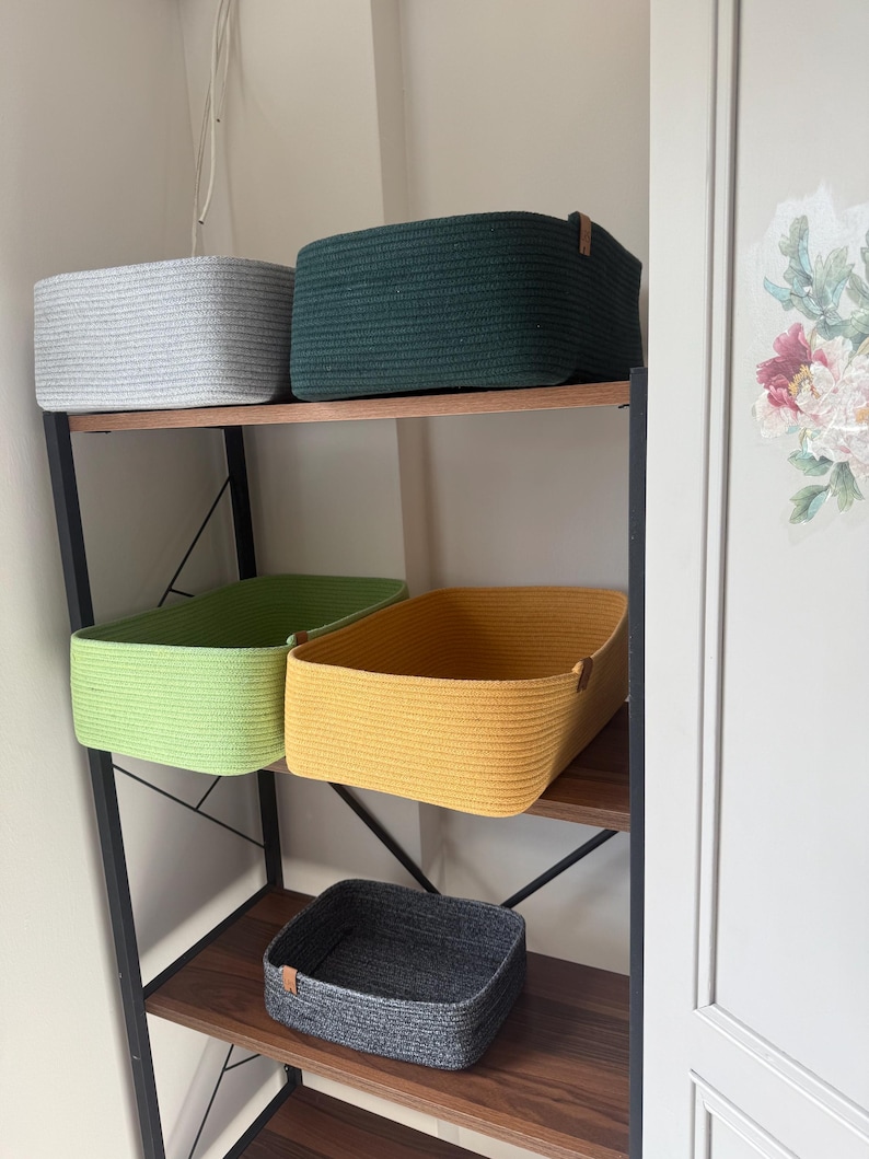 May include: Five rectangular storage baskets made of coiled rope, each featuring a small leather-like side tag. The baskets are colored light gray, dark green, lime green, mustard yellow, and charcoal gray, arranged on a dark wood shelving unit.