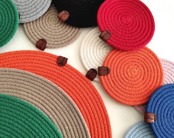 May include: A collection of colorful, circular coasters made from woven rope. The coasters are in various colors, including green, gray, black, red, orange, white, pink, and blue. Each coaster has a small, brown leather tag attached.