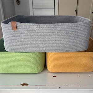 May include: Three rectangular storage baskets made of coiled rope. The top basket is light gray with a small brown leather tag embossed with the letters LRS. The bottom left basket is lime green, and the bottom right basket is mustard yellow.