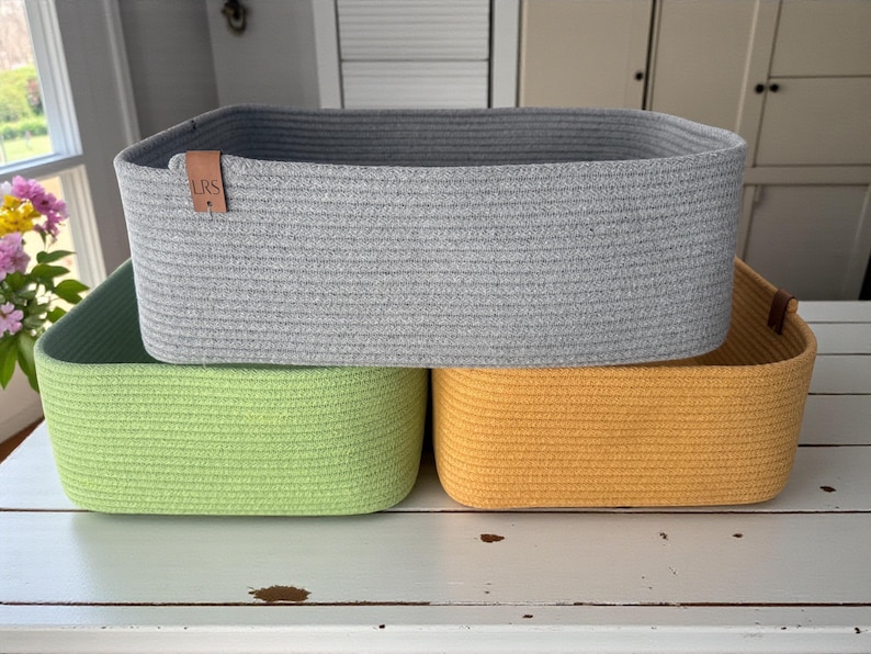 May include: Three rectangular storage baskets made of coiled rope. The top basket is light gray with a small brown leather tag embossed with the letters LRS. The bottom left basket is lime green, and the bottom right basket is mustard yellow.