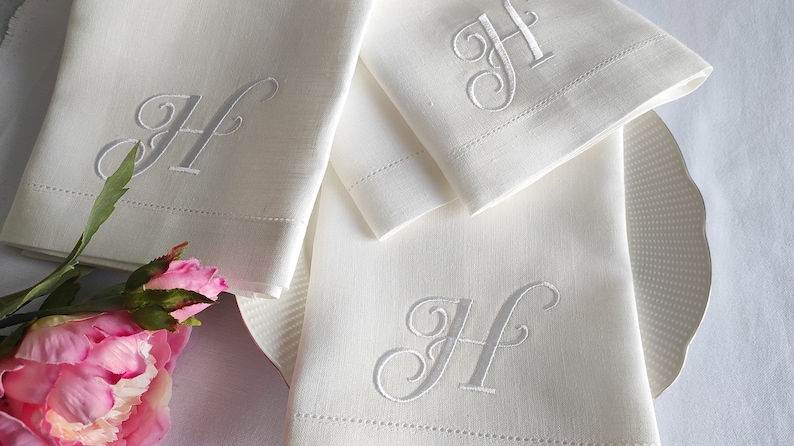 May include: Three white linen napkins with a silver embroidered letter "H" on each. The napkins are folded and arranged on a white surface with a pink flower.