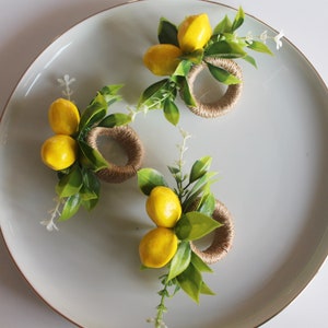 Pu&ograve; includere: Tre portatovaglioli decorativi con accenti di limone giallo e foglie verdi disposti su un piatto grigio chiaro. I portatovaglioli sono realizzati in spago intrecciato.