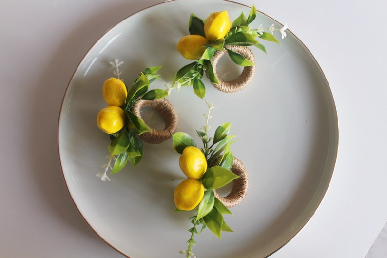 Pu&ograve; includere: Tre portatovaglioli decorativi con accenti di limone giallo e foglie verdi disposti su un piatto grigio chiaro. I portatovaglioli sono realizzati in spago intrecciato.