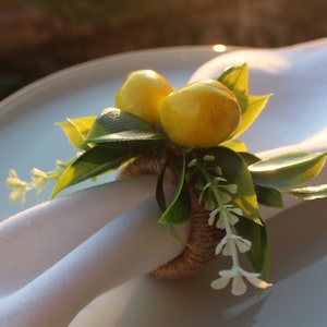 Pu&ograve; includere: Un portatovagliolo decorativo con limoni gialli, foglie verdi e piccoli fiori bianchi. L'anello &egrave; fatto di spago intrecciato e tiene un tovagliolo di lino bianco su un piatto bianco.