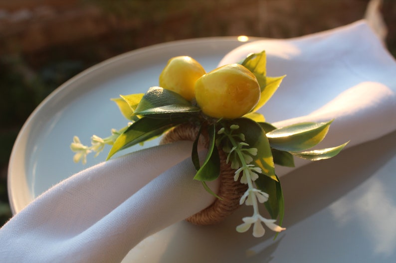 Pu&ograve; includere: Un portatovagliolo decorativo con limoni gialli, foglie verdi e piccoli fiori bianchi. L'anello &egrave; fatto di spago intrecciato e tiene un tovagliolo di lino bianco su un piatto bianco.