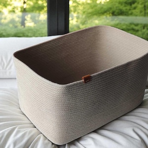 May include: A beige rectangular storage basket with a brown leather tag. The basket is made of woven material and sits on a white bed with pillows. The background shows a window with a view of green trees.