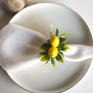 Pu&ograve; includere: Un tovagliolo di lino bianco, piegato e posto su un piatto bianco con bordo dorato, &egrave; decorato con un anello portatovagliolo a tema limone. L'anello presenta due limoni gialli, foglie verdi e piccoli fiori bianchi.