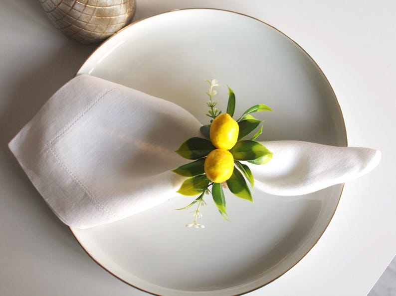Pu&ograve; includere: Un tovagliolo di lino bianco, piegato e posto su un piatto bianco con bordo dorato, &egrave; decorato con un anello portatovagliolo a tema limone. L'anello presenta due limoni gialli, foglie verdi e piccoli fiori bianchi.