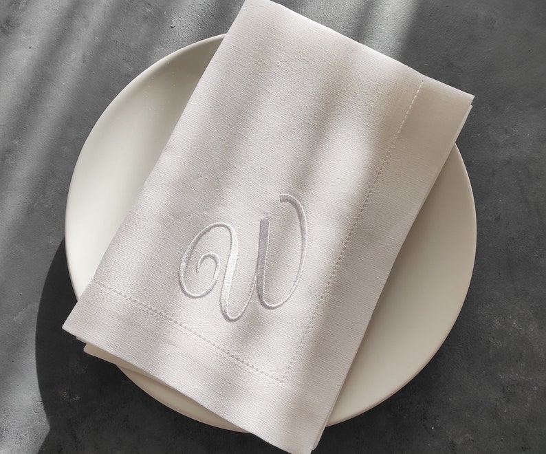 May include: White linen napkin with a white embroidered monogram "AW" on a white plate.