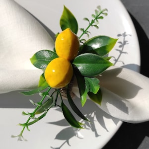 May include: A white napkin ring with a faux lemon and green leaves. The ring is made of burlap and has a rustic look.
