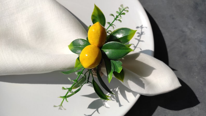 May include: A white napkin ring with a faux lemon and green leaves. The ring is made of burlap and has a rustic look.