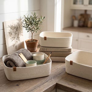 May include: Three rectangular off-white woven rope storage baskets with brown faux leather tabs. One basket contains a folded gray knit textile, a small gold metal ruler, and two small ceramic bowls. The baskets feature a thick, coiled rope texture.