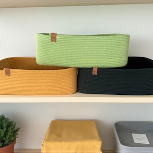 May include: Three rectangular storage baskets made of coiled rope in mustard yellow, lime green, and black. Each basket features a small brown leather tag with the letters LRS embossed on the front. The baskets are arranged on a shelf.