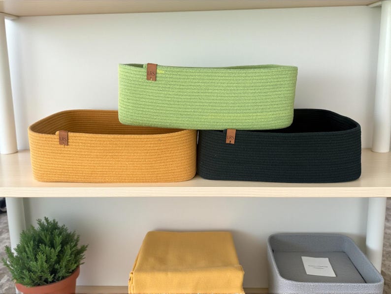 May include: Three rectangular storage baskets made of coiled rope in mustard yellow, lime green, and black. Each basket features a small brown leather tag with the letters LRS embossed on the front. The baskets are arranged on a shelf.