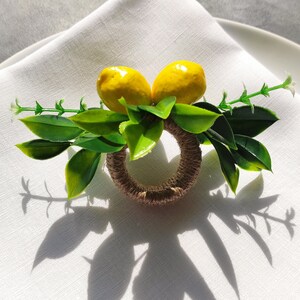 May include: A brown and beige napkin ring with two yellow lemons and green leaves. The ring is on a white linen napkin.