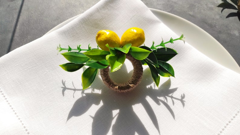 May include: A brown and beige napkin ring with two yellow lemons and green leaves. The ring is on a white linen napkin.