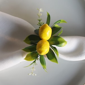 Pu&ograve; includere: Un portatovagliolo decorativo con due repliche di limoni gialli, foglie verdi e piccoli fiori bianchi. L'anello &egrave; posto su un tovagliolo bianco e un piatto bianco, creando un'estetica fresca ed estiva. Ideale per l'apparecchiatura della tavola.