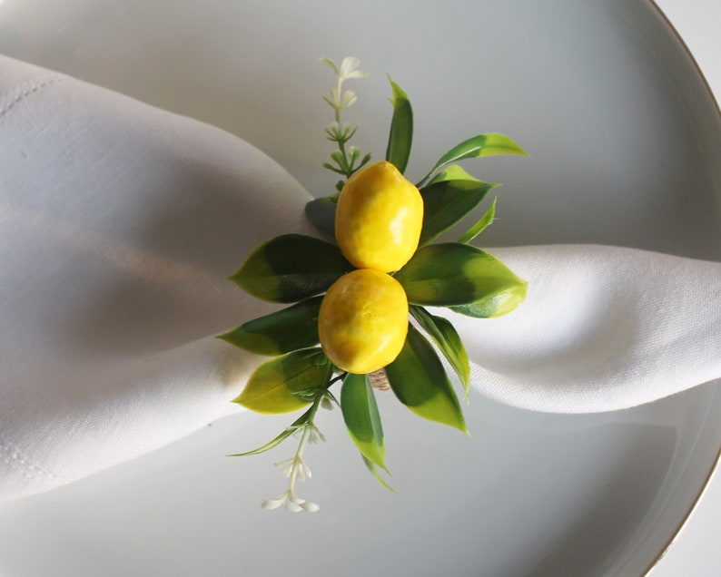 Pu&ograve; includere: Un portatovagliolo decorativo con due repliche di limoni gialli, foglie verdi e piccoli fiori bianchi. L'anello &egrave; posto su un tovagliolo bianco e un piatto bianco, creando un'estetica fresca ed estiva. Ideale per l'apparecchiatura della tavola.