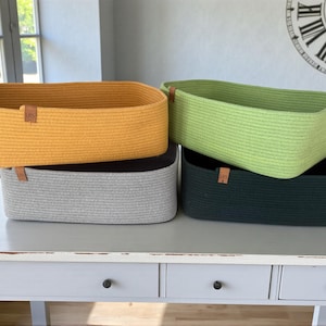 May include: Four rectangular woven storage baskets in shades of yellow, green, gray, and dark green. Each basket has a rectangular shape and a small leather tag. The baskets are stacked on a white table.