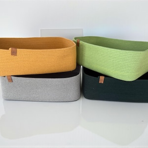 May include: Four rectangular storage baskets made of coiled rope in mustard yellow, light green, gray, and dark green. Each basket features a small, square brown leather-like tag on the side with the letters LRS embossed in a dark brown color.