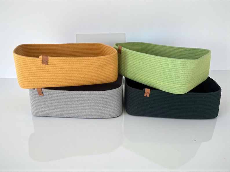 May include: Four rectangular storage baskets made of coiled rope in mustard yellow, light green, gray, and dark green. Each basket features a small, square brown leather-like tag on the side with the letters LRS embossed in a dark brown color.