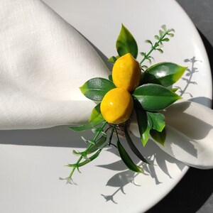May include: A white linen napkin with a lemon-themed napkin ring. The ring features two yellow lemons with green leaves.