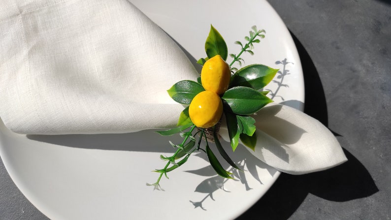 May include: A white linen napkin with a lemon-themed napkin ring. The ring features two yellow lemons with green leaves.