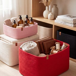 May include: A rectangular red woven rope storage basket with a small brown leather-like tab, containing a folded light-colored textile, two brown leather-bound notebooks, and several wooden spools with thread, sitting on a light wood surface.