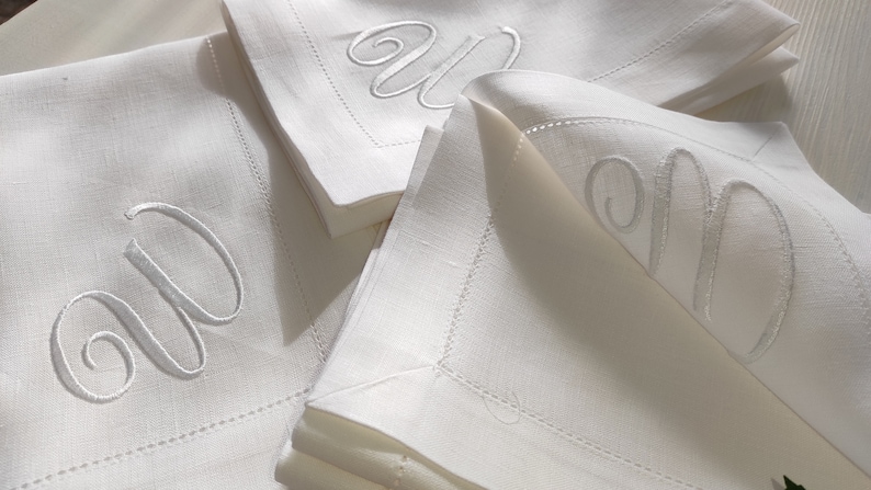 May include: Three white linen napkins with a white embroidered letter "W" on each. The napkins are folded and stacked on top of each other.