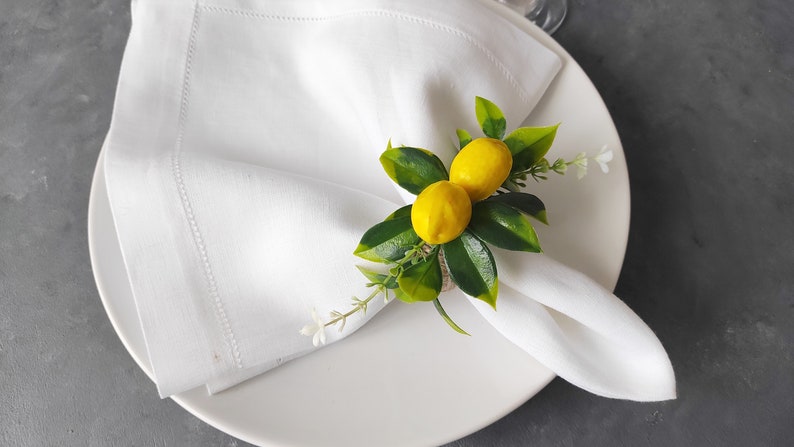 May include: White linen napkin with a lemon napkin ring. The ring is made of burlap and has two small yellow lemons with green leaves.