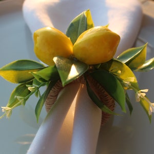 Pu&ograve; includere: Un portatovagliolo decorativo con due limoni gialli, foglie verdi e piccoli fiori bianchi. L'anello &egrave; fatto di spago intrecciato e tiene un tovagliolo bianco. Adatto per l'arredamento della tavola.