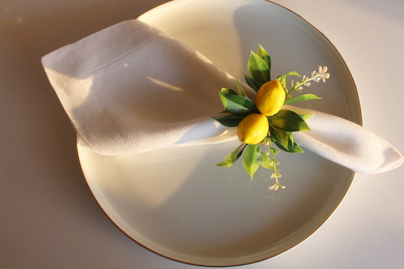 Pu&ograve; includere: Un tovagliolo di lino bianco, piegato e posto su un piatto bianco con bordo dorato. Il tovagliolo &egrave; decorato con un anello portatovagliolo a tema limone con due limoni gialli, foglie verdi e piccoli fiori bianchi. L'ambiente &egrave; luminoso.