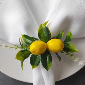 May include: A white napkin ring with two yellow lemons and green leaves. The lemons are surrounded by green leaves and white flowers.