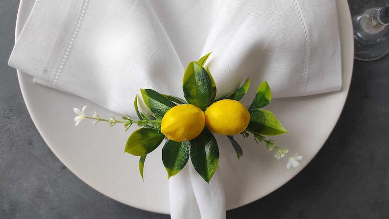 May include: A white napkin ring with two yellow lemons and green leaves. The lemons are surrounded by green leaves and white flowers.
