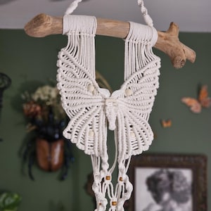 May include: A handmade macrame butterfly wall hanging. The butterfly is crafted from white cotton cord, with a detailed wing design. It is suspended from a natural driftwood branch. Decorative wooden beads add detail to the design. The background is a muted green.