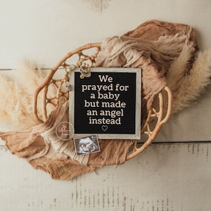 May include: A white framed letter board with black felt letters spelling "We prayed for a baby but made an angel instead" with a small heart at the end. The letter board is sitting in a woven basket with a tan and white fabric draped over it.