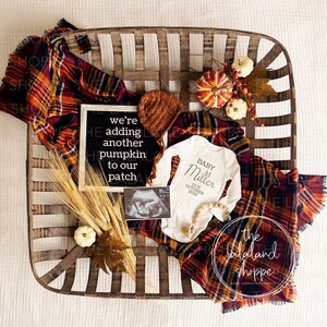 Könnte beinhalten: Herbstliche Babyankündigung mit einem Weidenkorb, einem karierten Schal und dekorativen Kürbissen. Die Ankündigung enthält einen Strampler, eine Strickmütze und ein Schild mit der Aufschrift "we're adding another pumpkin to our patch."