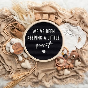 May include: A tan blanket with a wooden letter board that reads "We've been keeping a little secret" with a white heart. There are wooden blocks that spell "BABY", a white onesie that reads "Baby Parker", a wooden sign that reads "Due August 2024", a wooden bunny, and a wooden bead garland.