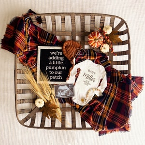 May include: A wooden basket filled with autumnal decor, including a plaid blanket, a pumpkin, and a sign that reads "we're adding a little pumpkin to our patch". A white onesie with the text "Baby Miller Due October 2023" is also in the basket.