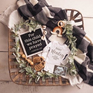 Puede incluir: Un body blanco con el texto "BABY Henley DUE OCTOBER 2023" en letras marrones, un tablero de letras negro con el texto "For this child we have prayed 1 SAMUEL 1:27", una cesta de madera con vegetación, un sonajero de madera, un juguete de crochet arcoíris y una pequeña imagen de ultrasonido.