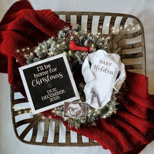 May include: A white onesie with the text "BABY Holden" on it, a black letter board with the text "I'll be home for Christmas" and a heart, "DUE DECEMBER 2023", a Christmas wreath with red berries and lights, and a red blanket.