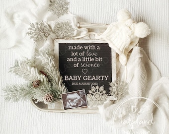 May include: A black chalkboard sign with white text that reads "made with a lot of love and a little bit of science" with a heart symbol. Below the text is "BABY GEARTY" and "DUE AUGUST 2023". The sign is surrounded by white fabric, a white knitted hat, a small pine branch, and a silver snowflake.