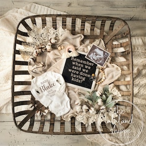 May include: A white onesie with the text "BABY Blake DUE OCTOBER 2023" in pink lettering, a black letter board with the text "Remember when we said we were done having kids?" and a small ultrasound image in a wooden basket with cotton balls, greenery, and a wooden bead necklace.