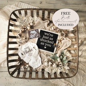 May include: A white onesie with the text "BABY Blake DUE AUGUST 2024" on it, a black letter board with the text "we're just as shocked as you", a white blanket, a wooden basket, a cotton ball garland, and a white cotton flower arrangement.