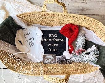 May include: A wicker basket filled with baby items, including a white onesie with the text "Baby Hines Due June 2022", a red Santa hat, a black chalkboard with the text "and then there were four", a white ultrasound image, and a sprig of greenery with white and red berries.