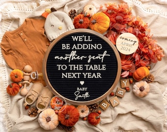 May include: A black letter board with the text "We'll be adding another seat to the table next year" and the words "Baby Smith" below. The board is surrounded by autumn-themed decorations, including pumpkins, pinecones, and autumn leaves. There is also a wooden circle with the text "coming April 2024" and wooden blocks that spell out "Baby".
